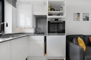 Une cuisine de taille réduite avec des meubles blancs, un four Bosch et un canapé gris agrémenté de coussins jaunes.