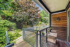 Un balcon doté d’une table et de chaises en bois, entouré d’une rambarde en métal, donnant sur un jardin avec des arbres et un mur en pierre.