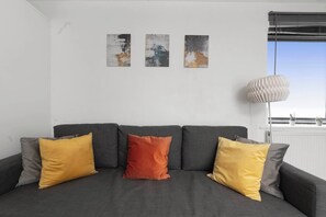 A grey sofa with three cushions in yellow and orange, a white radiator, and a window with blinds. - Hermitage Rd N4, Manor House | Garden Studio (London)