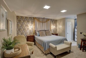 Ein Schlafzimmer mit einem großen Bett, zwei Nachttischen, einem Sessel, einem kleinen Tisch mit einer Pflanze und einer Tür zu einem anderen Raum.