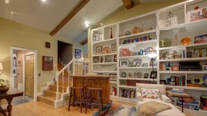 A cozy living room with a wooden staircase, a white sofa, and a bookshelf filled with books and decorative items. - Entire Spacious 4BR Home - close to downtown & minutes to trails! (Bentonville)