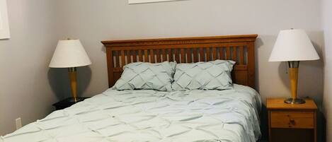 A bedroom with a wooden bed, two bedside tables with lamps, a window, and a wall-mounted light fixture.