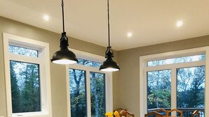 A modern kitchen with a central island, two pendant lights, a dining area with a wooden table and chairs, and large windows with a view of trees.