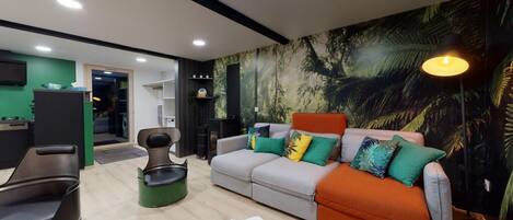 A modern living room with a sofa, a coffee table, and a green accent chair. There is a bookshelf and a desk with a computer.