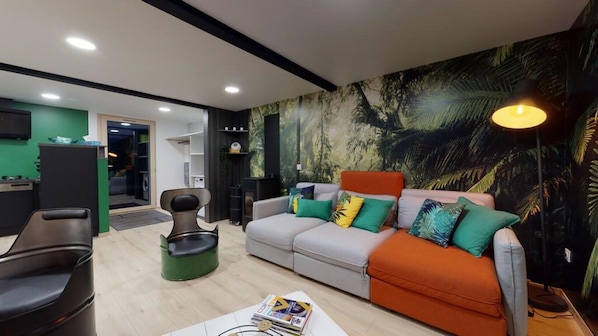 Una sala de estar moderna con un sofá, una mesa de centro y un sillón verde decorativo. Se observa una estantería y un escritorio con una computadora.