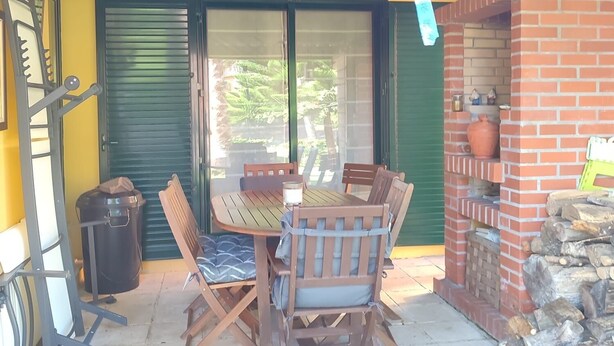 An outdoor patio with a wooden table and chairs, a trash bin, and a brick fireplace.