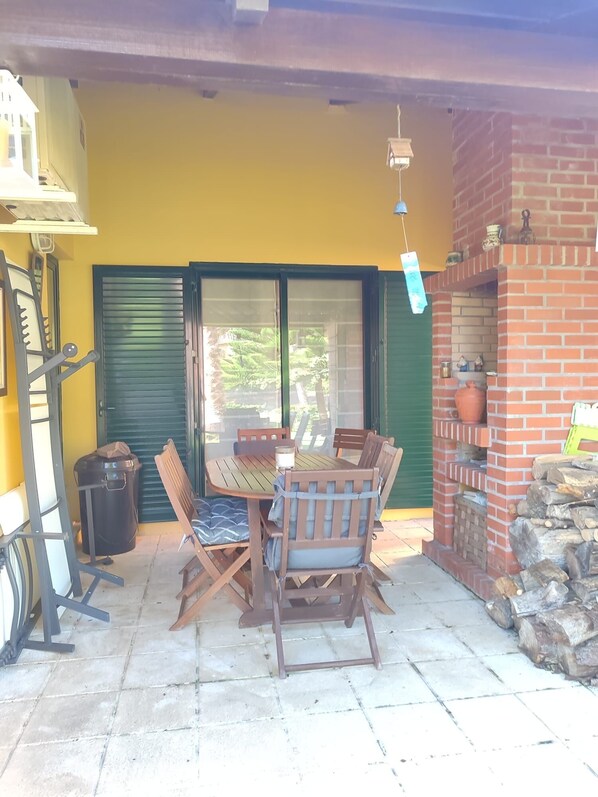 An outdoor patio with a wooden table and chairs, a trash bin, and a brick fireplace.