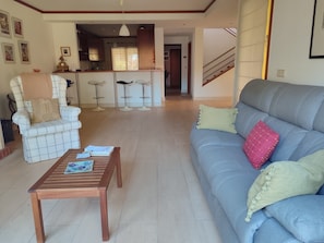 Una sala de estar con un sofá, un sillón y una mesa de centro. Se ve una cocina al fondo con una zona de comedor.