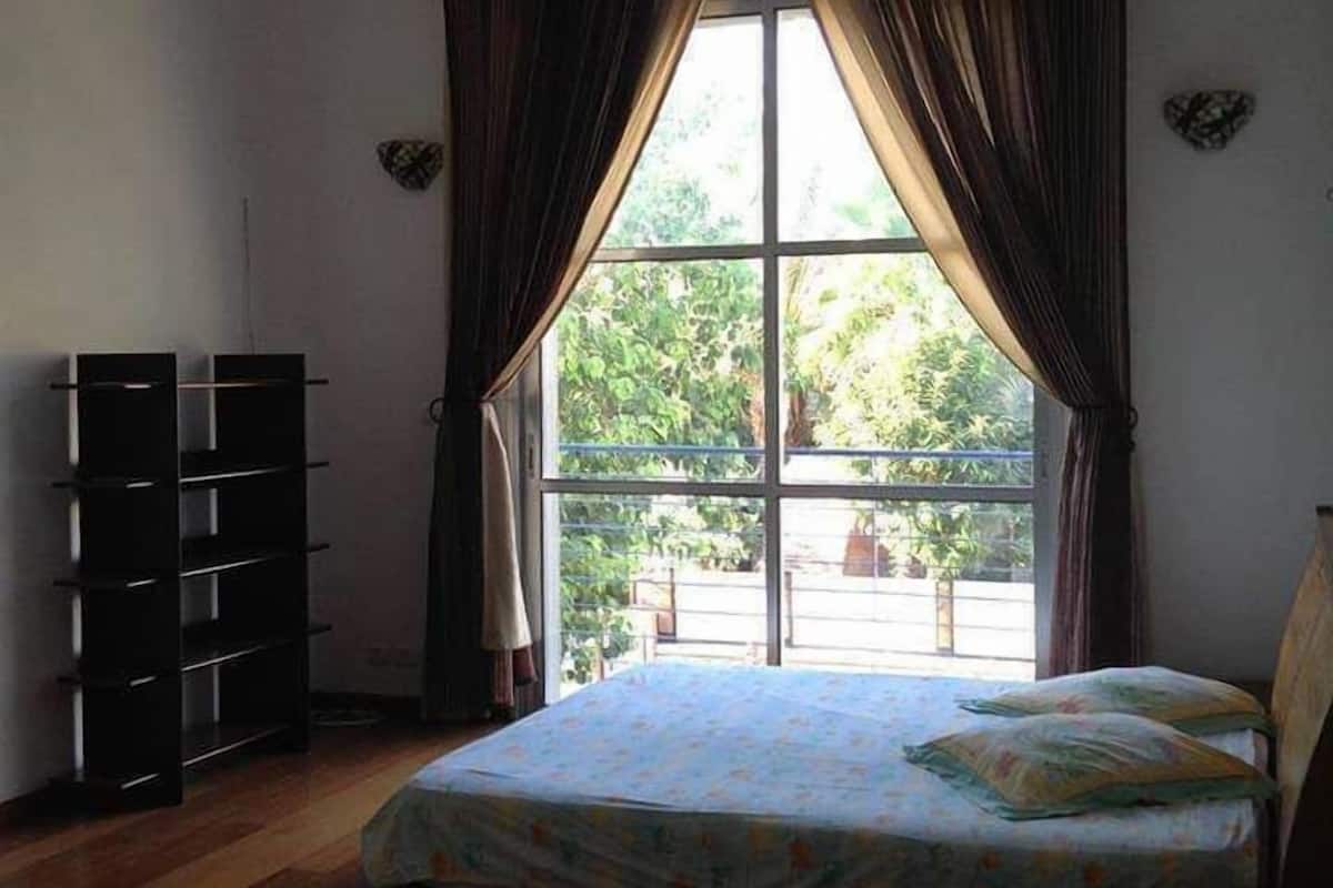 Een slaapkamer met een bed, een raam met gordijnen, een boekenkast en uitzicht op groen.
