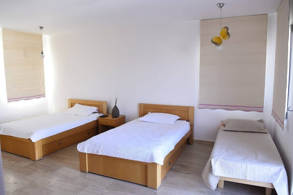 Een kamer met drie eenpersoonsbedden, houten hoofdborden, een nachtkastje met een vaas en een hanglamp.
