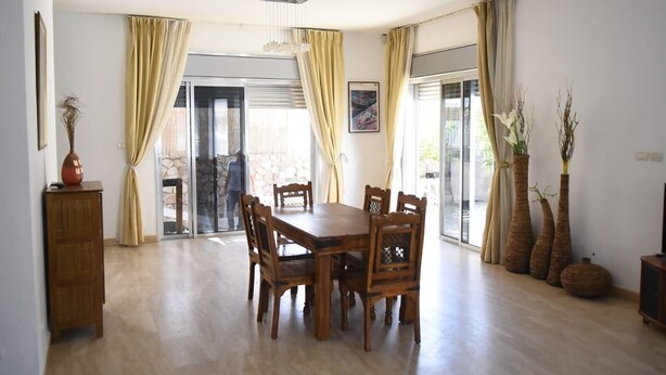 A dining area with a wooden table and chairs, a vase with dried plants, and a view through a glass door to an outdoor area. - Casa de la familia (Eilat)