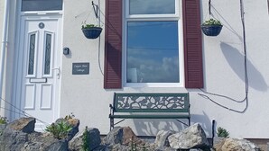 Une petite maison blanche avec deux fenêtres, des volets marron, une porte avec un numéro et un banc agrémenté de jardinières suspendues.