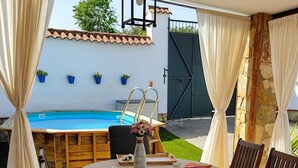 Un espace extérieur aménagé pour les repas, avec une table ronde pour quatre personnes, entourée de chaises en bois. On aperçoit une piscine à l’arrière-plan, et des plantes en pot sont disposées le long du mur.
