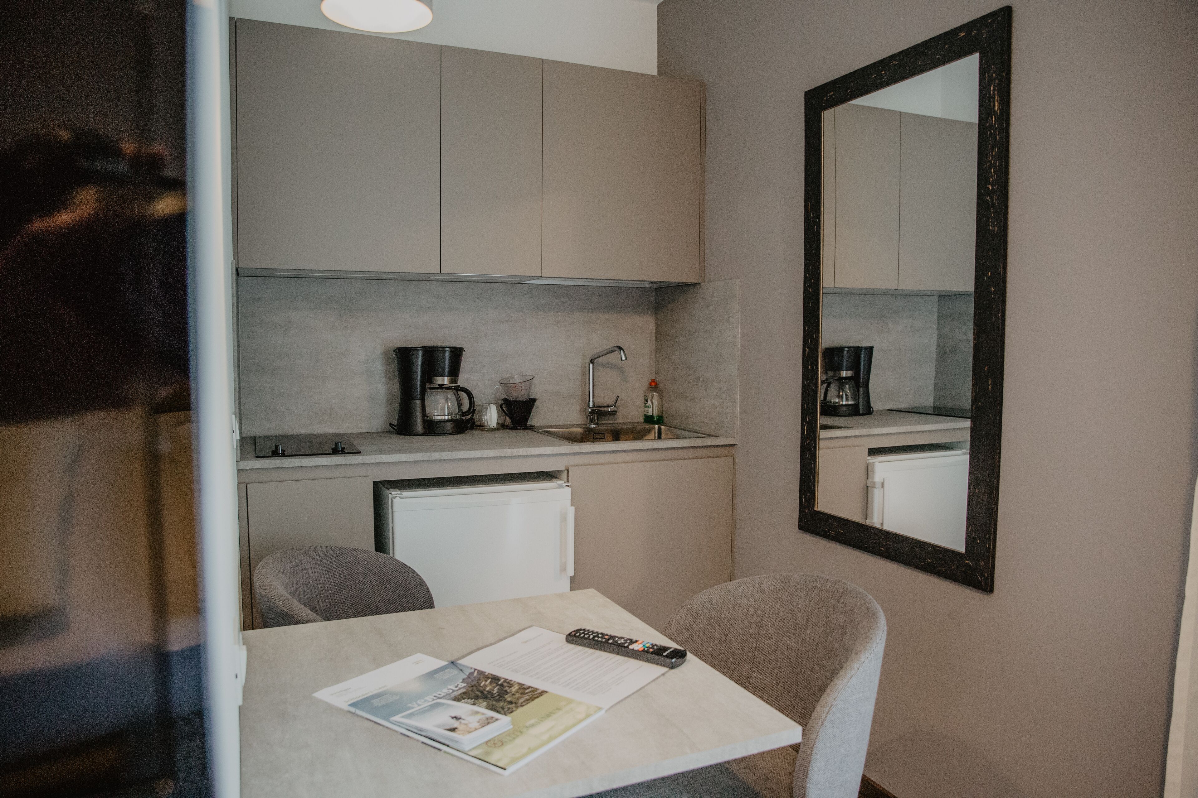 Una pequeña kitchenette con cafetera, hervidor y lavamanos. Hay una mesa con un control remoto y una revista. Un espejo está en la pared.