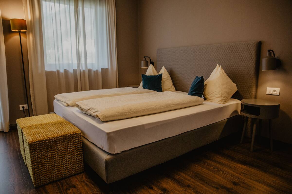 Una cama doble con cabecera, dos lámparas de noche, una mesita de noche y un taburete tejido.