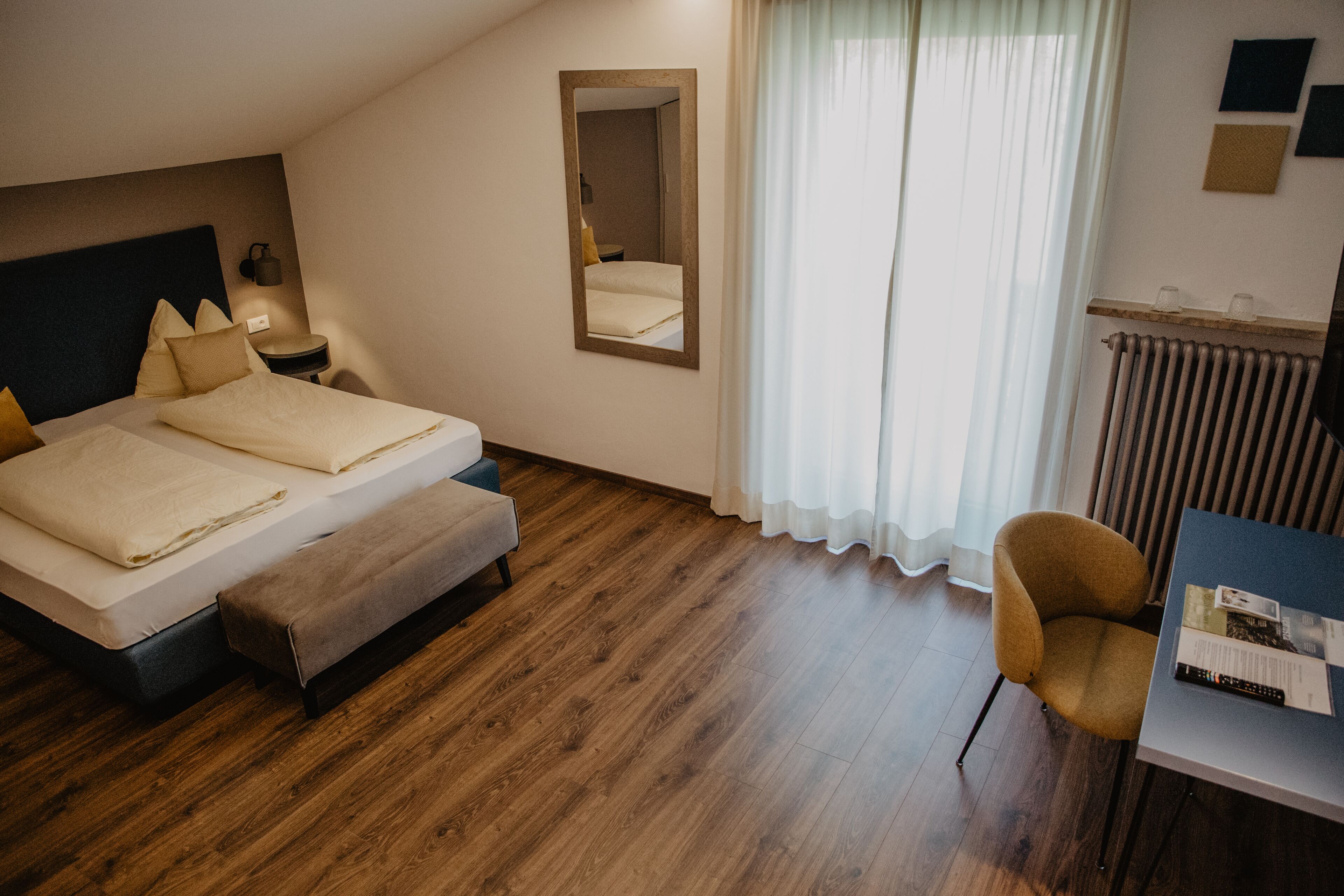 Una habitación de hotel con una cama, un banco, una silla, un espejo, una mesita de noche y un radiador.