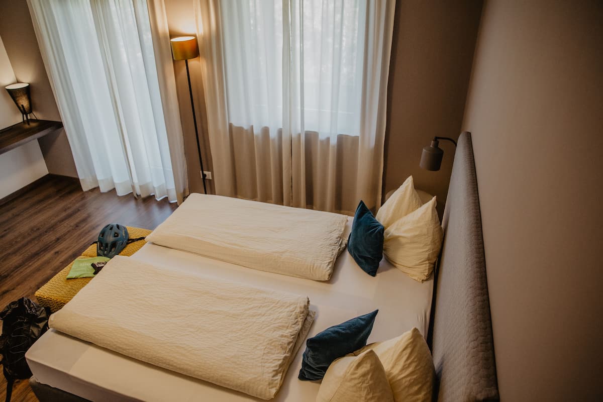 Habitación de hotel con cama, almohadas, cabecero, mesita de noche, lámpara y ventana con cortinas.