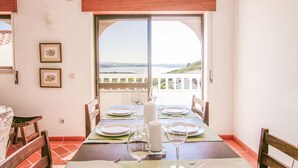 Ein Essbereich mit Tisch für zwei, Meerblick durch ein großes Fenster und ein Balkon mit Geländer.