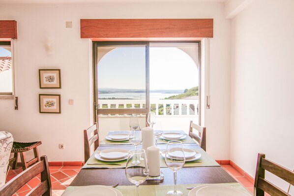A dining area with a table set for two, a view of the sea through a large window, and a balcony with a railing.