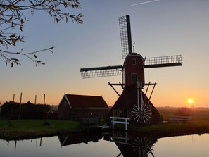 A traditional windmill with a red body and white accents, situated next to a body of water with a reflection, and a sunset in the background. - Lake house 74 luxury accommodation with iR Sauna (Warmond)