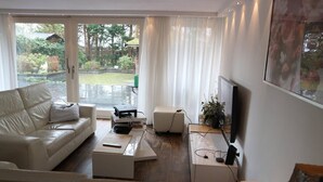 A modern living room with a white leather sofa, a coffee table, a TV, and a large window overlooking a garden. - Lake house 74 luxury accommodation with iR Sauna (Warmond)