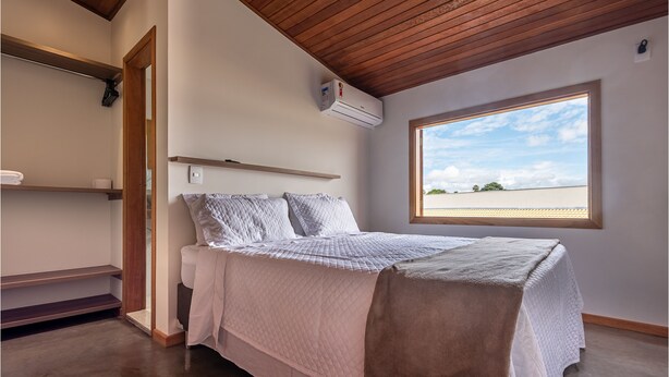 Een slaapkamer met een bed, een raam, een houten plafond en een luchtverkoeler aan de muur.