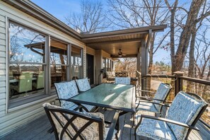 A covered outdoor patio with a glass table and chairs, surrounded by trees and a wooden deck. - The Perch on Beaver Lake (Rogers)