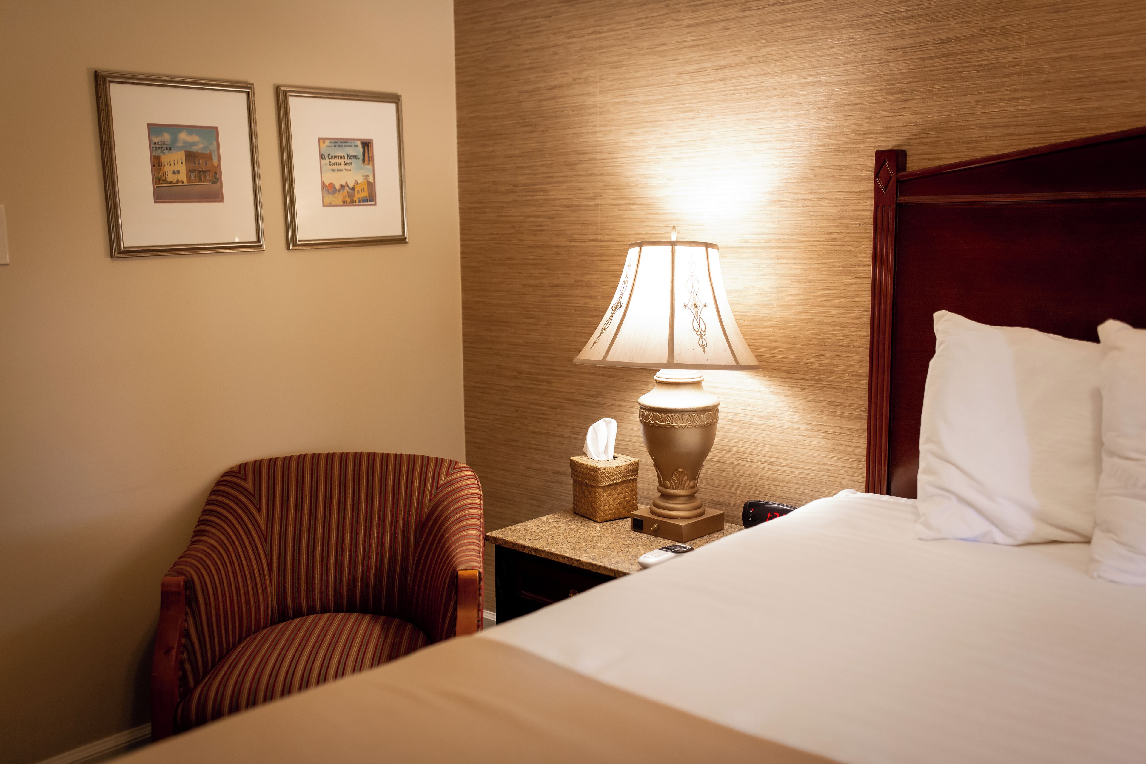 A hotel room with a bed, a chair, a nightstand with a lamp, and two framed pictures on the wall.