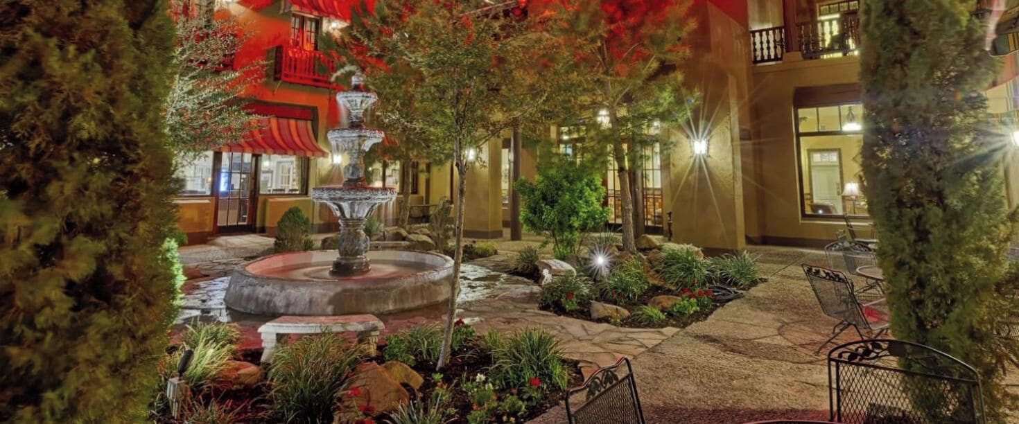 A courtyard with a central fountain, surrounded by potted plants and trees, and a building with balconies in the background.