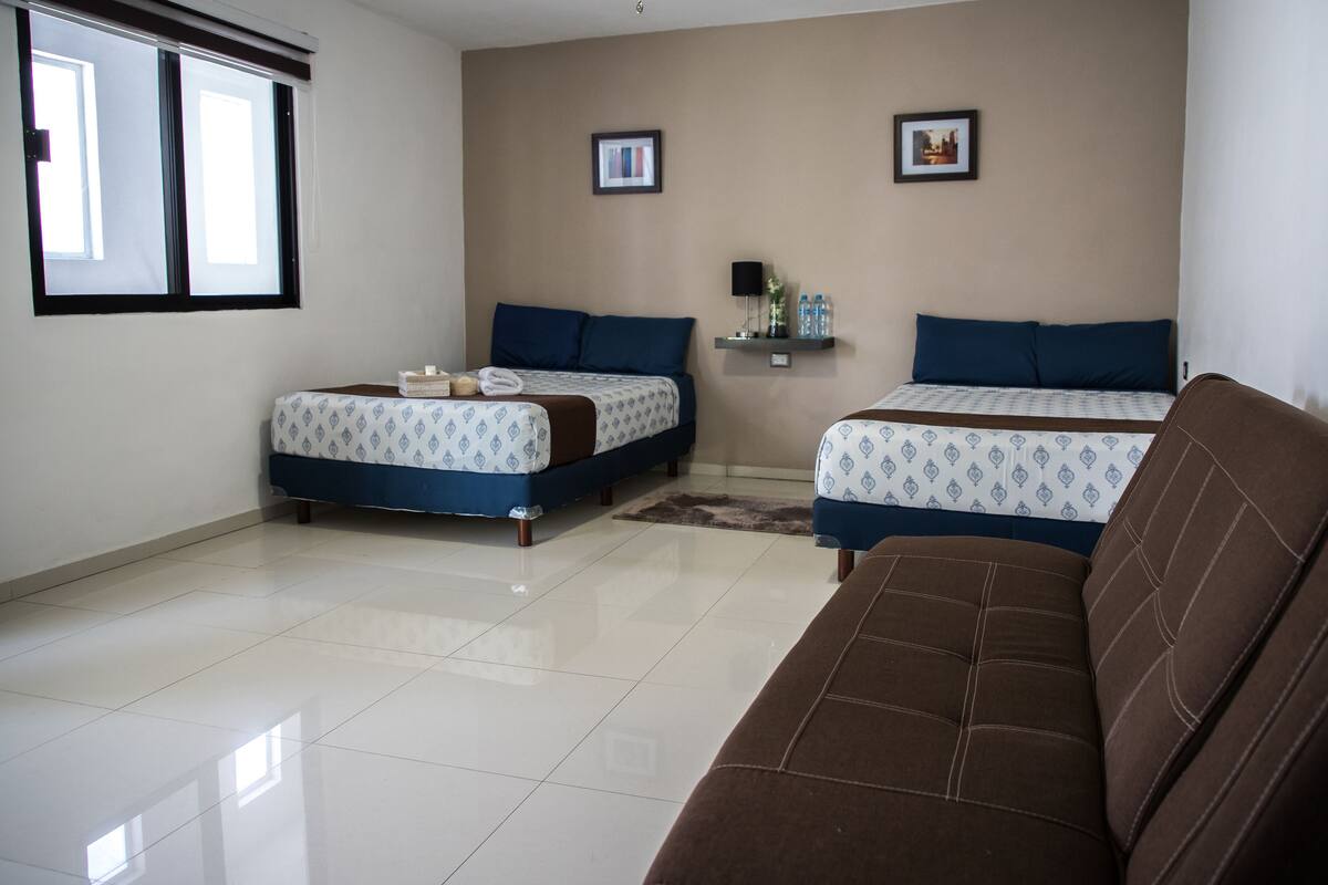 A hotel room with two beds, a brown sofa, a tiled floor, and two framed pictures on the wall.