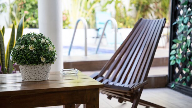 A wooden table with a decorative pot and a glass on it, a wooden lounge chair, and a pool area in the background. - Margarett Hotel (Valladolid)