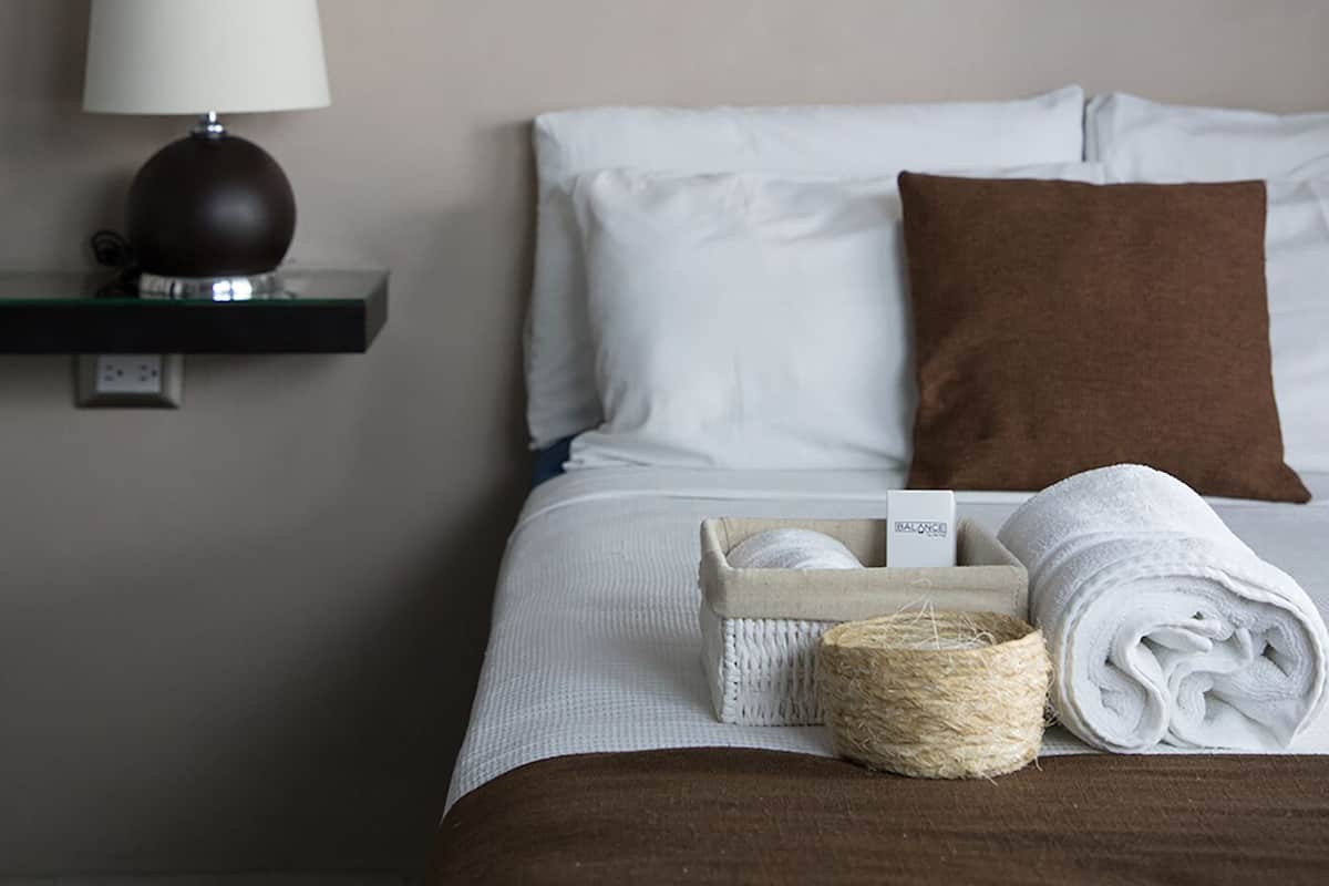 A neatly made bed with white and brown bedding, a bedside lamp, a framed picture on the wall, and a small basket with towels and a spool of twine.