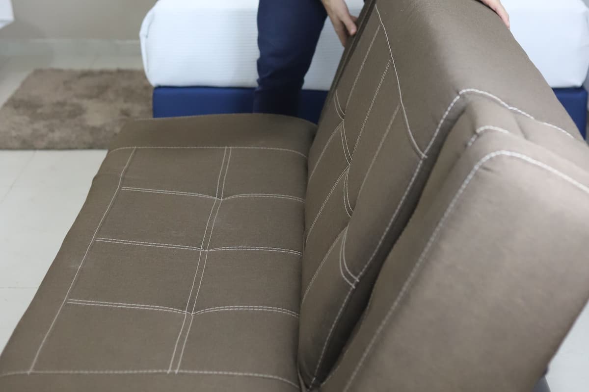 A person adjusting a brown sofa in a room with a bed and a bedside table with a lamp.