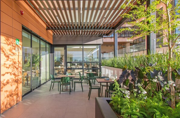 An outdoor seating area with tables and chairs, a wooden pergola, and a view of the cityscape through large windows.