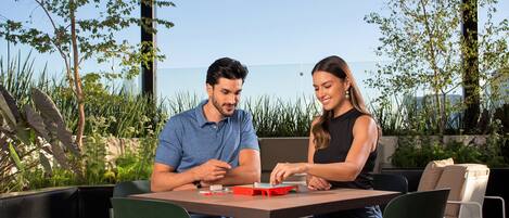 Two people seated at an outdoor table with a view of the sea, surrounded by greenery.
