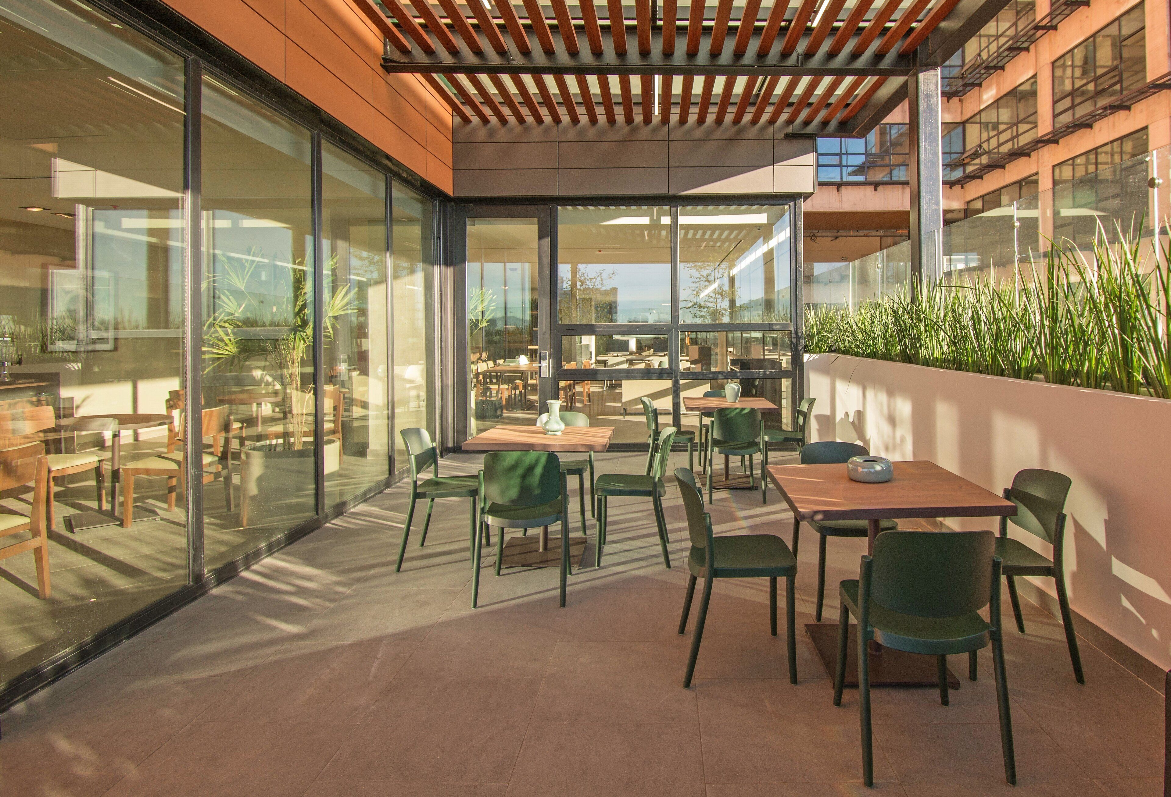 An outdoor patio area with glass walls, wooden tables, and green chairs.