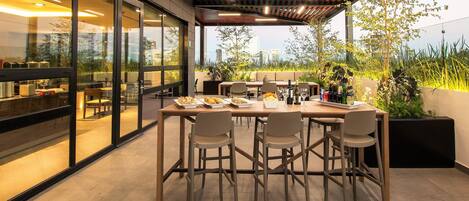 Un espace de restauration en plein air au design moderne, avec une table et des chaises en bois, entouré de murs en verre et offrant une vue sur le paysage urbain.