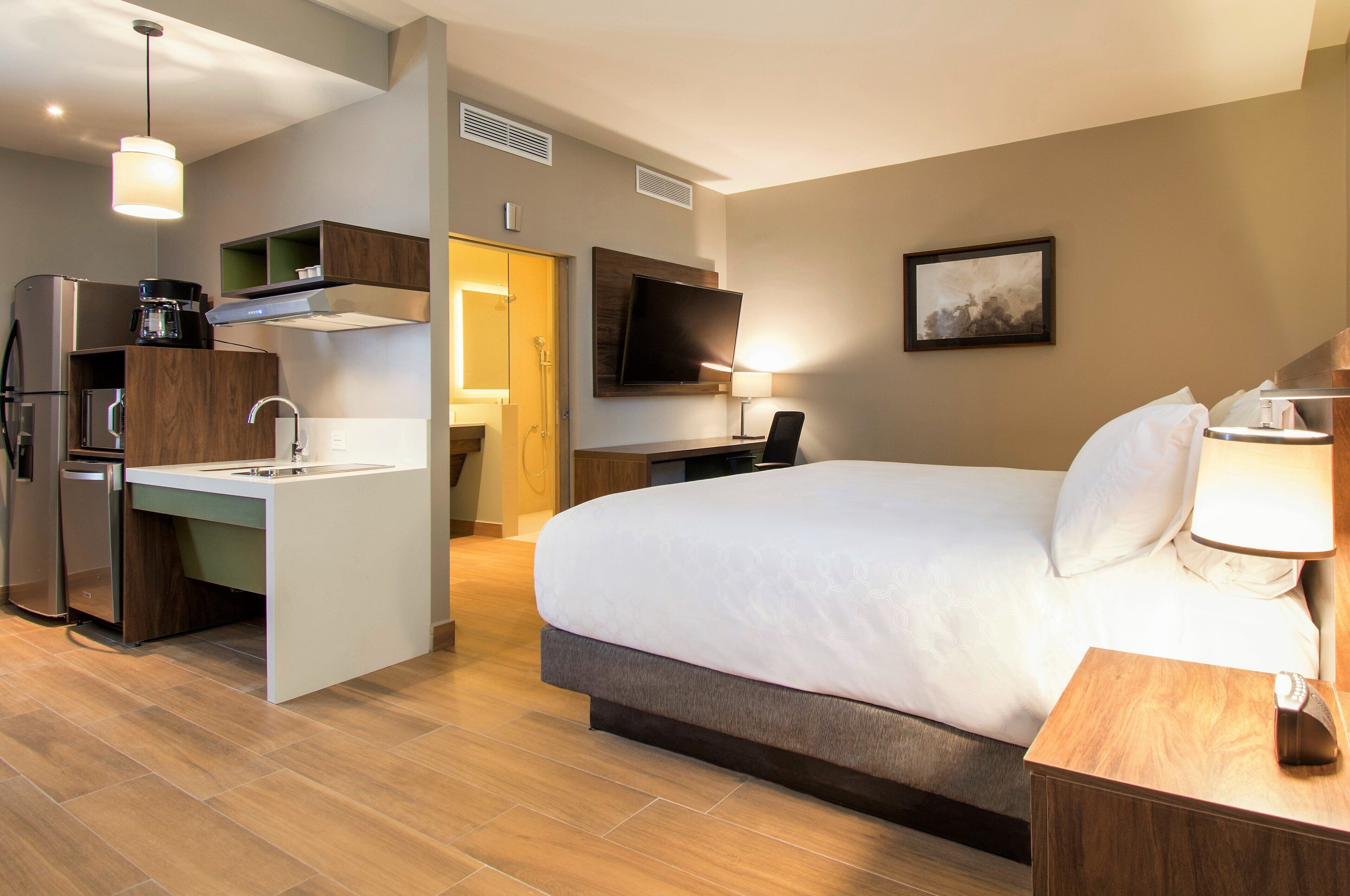 A hotel room with a large bed, a desk with a chair, a kitchenette, a TV, and a bathroom.