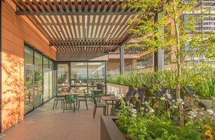 Un área de café al aire libre con mesas y sillas, rodeada de follaje y una pérgola de madera.