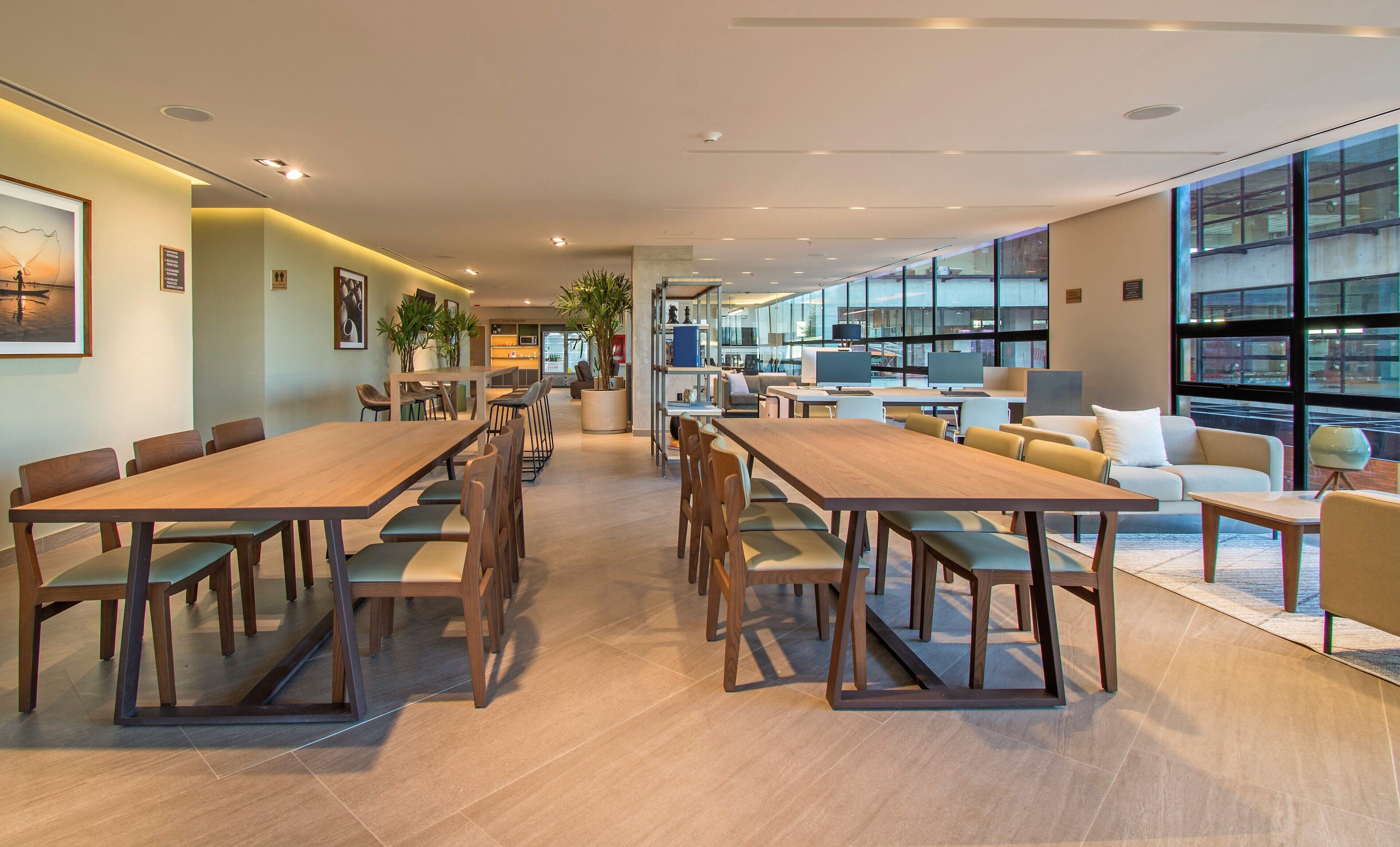 Un vaste espace de salle à manger moderne, doté de longues tables et de chaises en bois, d’un coin cuisine équipé d’appareils électroménagers modernes et de grandes fenêtres offrant une vue sur la ville.