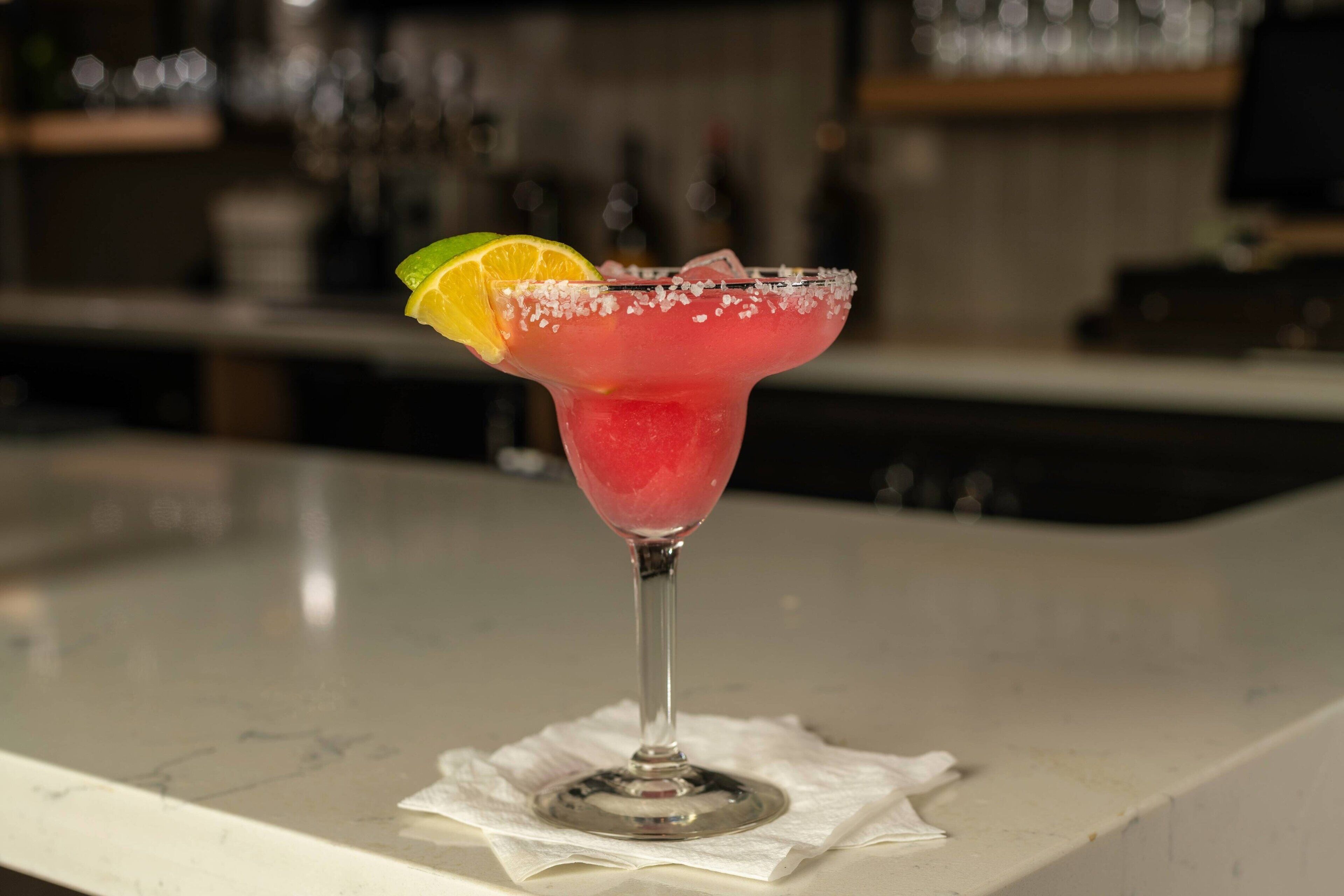 A bar setting with a pink cocktail in a glass with a salted rim, garnished with a lime slice.