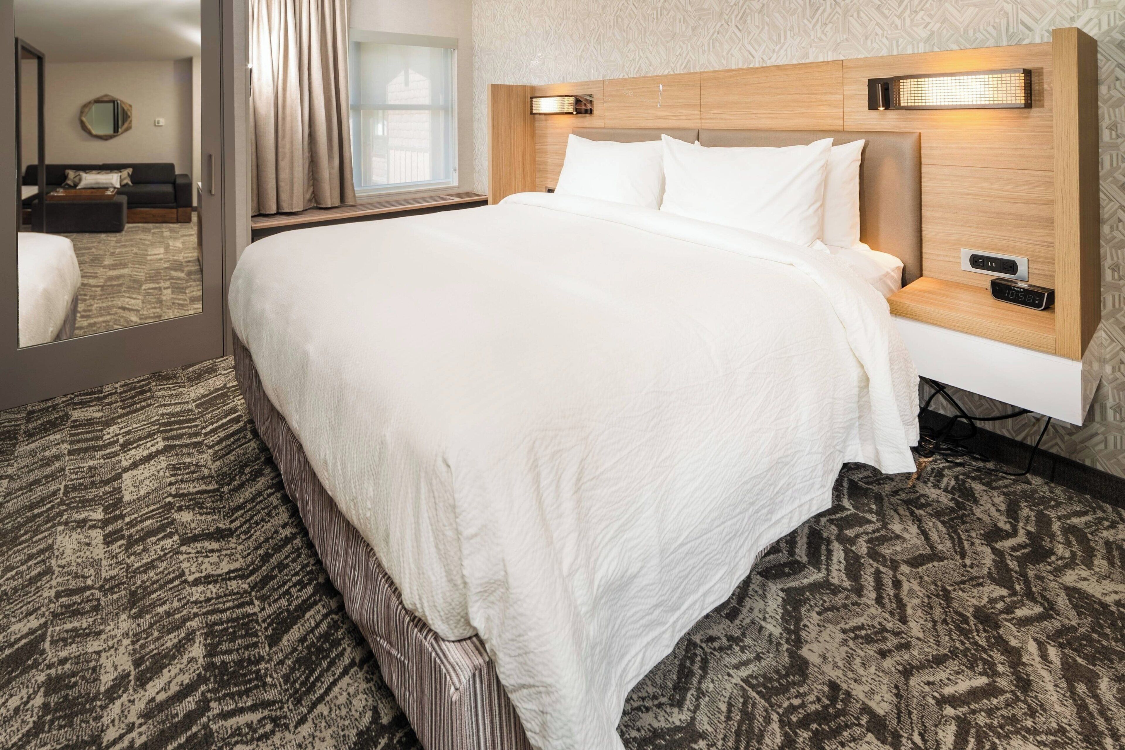 Una habitación de hotel con una cama grande, una mesita de noche con reloj digital, un cabecero con iluminación integrada y una alfombra estampada.