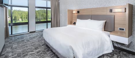 A modern hotel room with a large bed, a TV mounted on the wall, and a view of a forest through large windows.