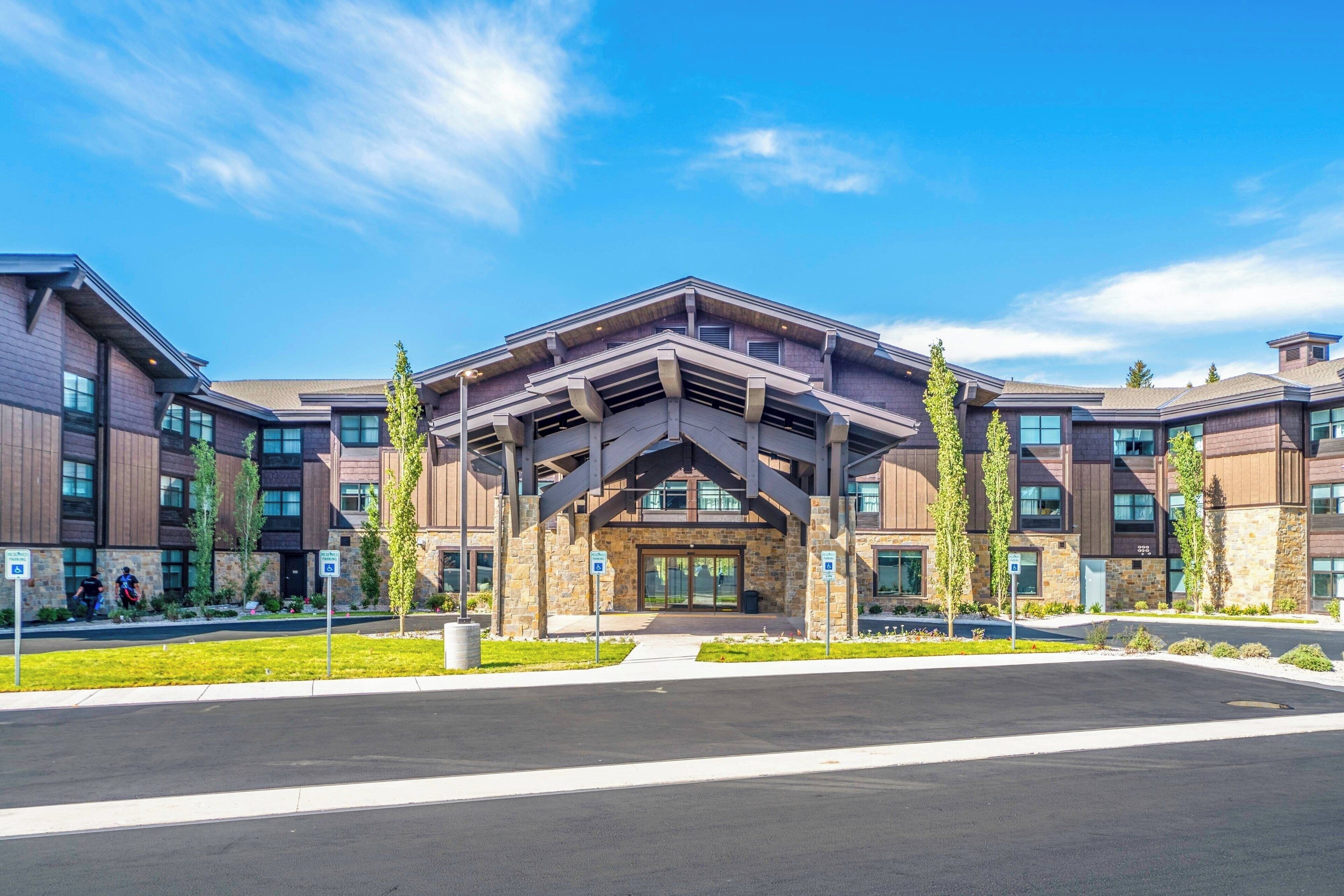 A modern building with a central entrance, multiple windows, and a parking lot in front.