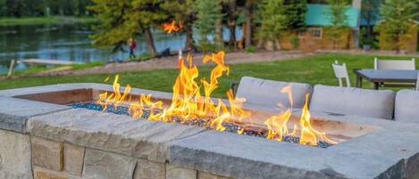 Un foyer en pierre avec des flammes, entouré de meubles d’extérieur, le tout installé sur un terrain donnant sur un lac, avec des arbres et une maison en arrière-plan.