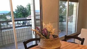 Taula de dinar amb cadires, cistella teixida amb flors i grans finestres que donen a un balcó i edificis.