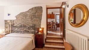 Una camera da letto con parete in pietra, un letto con testiera, un comodino con lampada e vista sulla cucina attraverso una porta aperta.