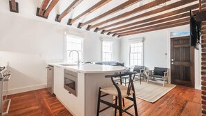 Een ruime keuken met een groot eiland, houten balken in het plafond en uitzicht op de woonkamer met een bank en stoelen.