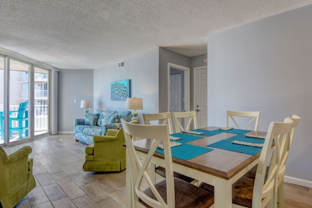 Un comedor con una mesa de madera y sillas blancas, un sofá azul y un sillón verde en una habitación con paredes azul claro y un ventanal grande.