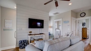 Un salon moderne avec un téléviseur à écran plat, des canapés gris, un ventilateur de plafond et un plancher en bois.